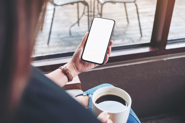 donna che tiene telefono mobile nero con schermo desktop vuoto mentre beve il caffè nella caffetteria