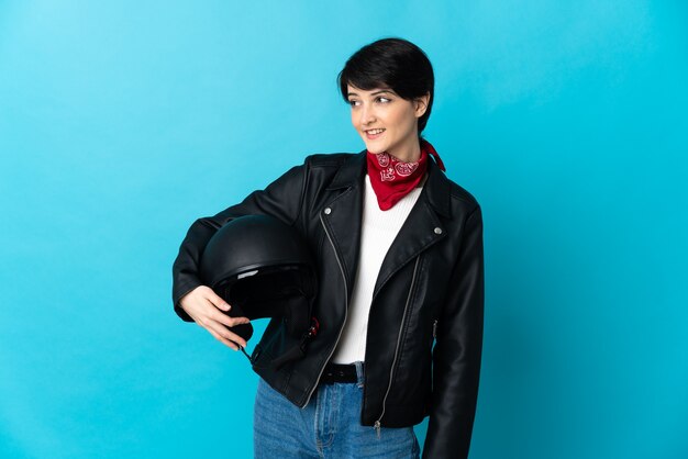 Donna che tiene un casco del motociclo isolato sulla parete blu che osserva al lato e sorridente