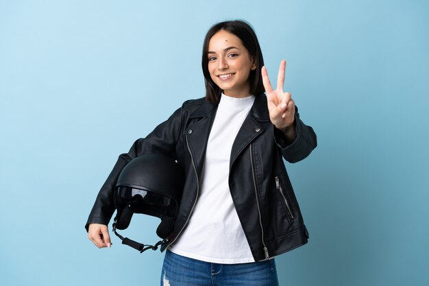 Donna che tiene un casco del motociclo isolato sulla parete blu che sorride e che mostra il segno di vittoria