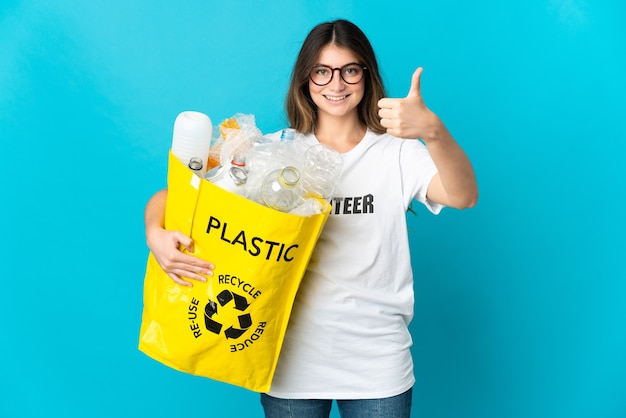 Donna che tiene una borsa piena di bottiglie da riciclare isolata sull'azzurro con i pollici in su perché è successo qualcosa di buono