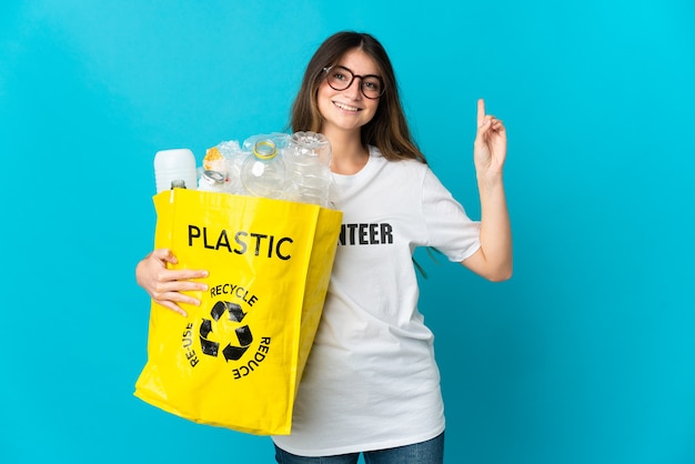 Donna che tiene una borsa piena di bottiglie da riciclare isolata sulla rappresentazione blu e alzando un dito in segno del meglio