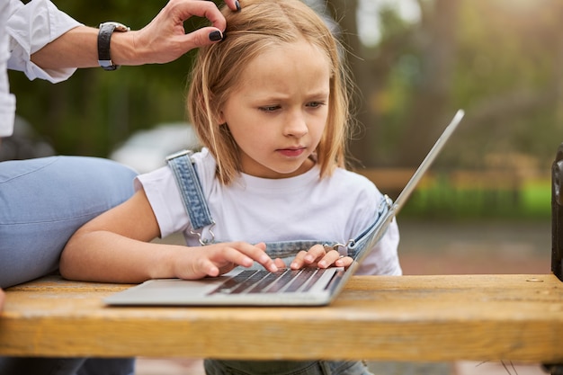 donna che tocca i capelli di una graziosa figlia affascinante all'aperto mentre una bambina guarda lo schermo del laptop