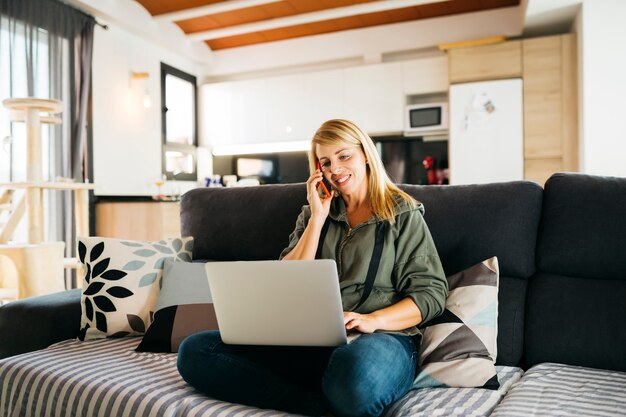 Donna che usa un laptop e conversa su uno smartphone rosso su un divano