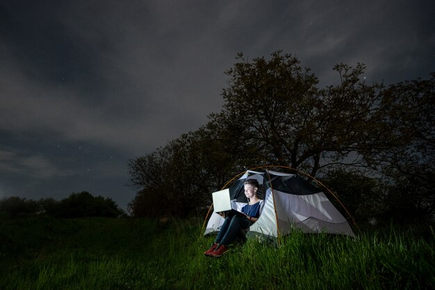 Donna che utilizza il suo computer portatile nel campeggio di notte. Femmina che si siede nella tenda sotto gli alberi e il cielo notturno