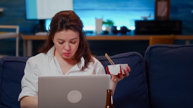 Donna che utilizza laptop per lavoro e mangia cibo asiatico a casa sul divano. Giovane adulto che si gode un pasto cinese da asporto mentre lavora su un dispositivo moderno e guarda la televisione in soggiorno