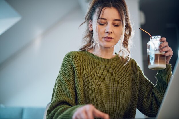 Donna che utilizza un computer portatile e beve caffè con cannuccia ecologica a casa