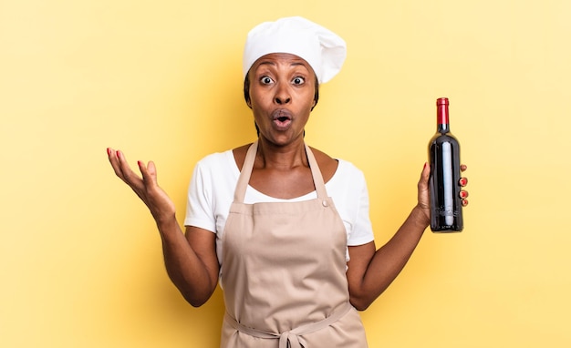 Donna chef afro nera a bocca aperta e stupita, scioccata e stupita da un'incredibile sorpresa. concetto di bottiglia di vino