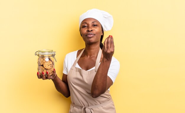 Donna chef afro nera che fa un gesto capice o denaro, dicendoti di pagare i tuoi debiti!. concetto di biscotti
