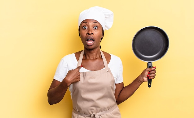 Donna chef afro nera che sembra scioccata e sorpresa con la bocca spalancata, che indica se stessa