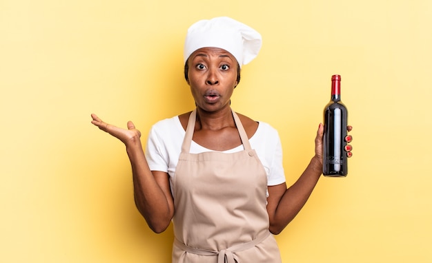 donna chef afro nera che sembra sorpresa e scioccata, con la mascella caduta in possesso di un oggetto con una mano aperta sul lato. concetto di bottiglia di vino