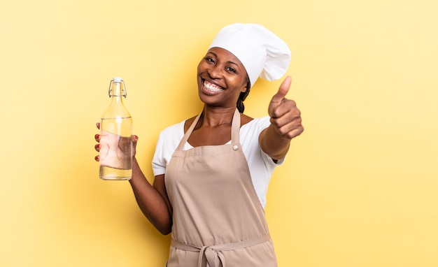 Donna chef afro nera che si sente orgogliosa, spensierata, sicura di sé e felice, sorridendo positivamente con il pollice in alto tenendo in mano una bottiglia d'acqua