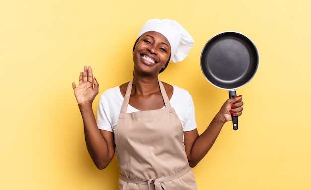 Donna chef afro nera che sorride allegramente e allegramente, agitando la mano, accogliendoti e salutandoti, o salutandoti