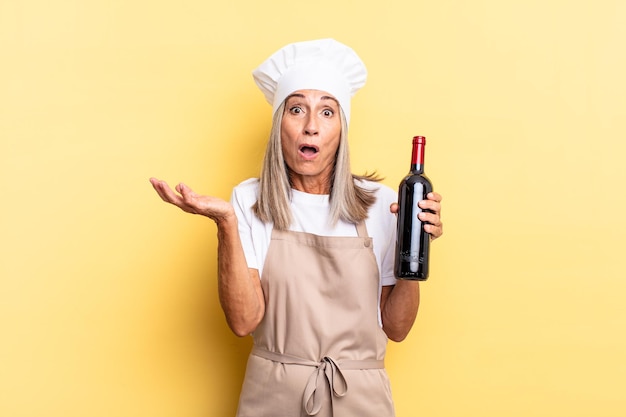Donna chef di mezza età a bocca aperta e stupita, scioccata e stupita con un'incredibile sorpresa con in mano una bottiglia di vino