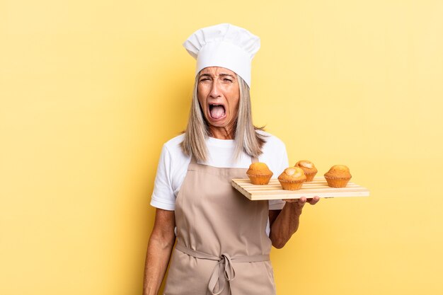 Donna chef di mezza età che grida in modo aggressivo, sembra molto arrabbiata, frustrata, indignata o infastidita, urla di no e tiene in mano un vassoio di muffin