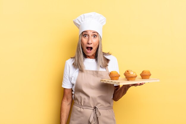 Donna chef di mezza età che sembra molto scioccata o sorpresa che fissa con la bocca aperta dicendo wow e tenendo un vassoio per muffin