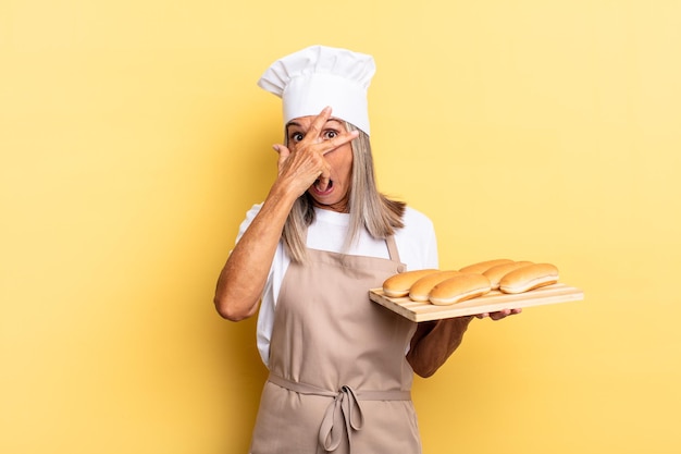 Donna chef di mezza età che sembra scioccata, spaventata o terrorizzata, coprendo il viso con la mano e sbirciando tra le dita e tenendo un vassoio per il pane