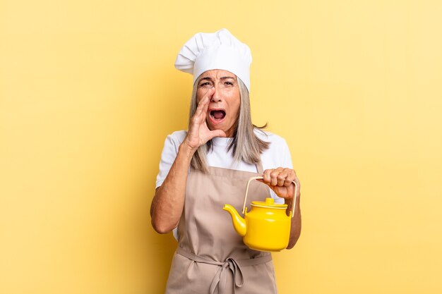 Donna chef di mezza età che si sente felice, eccitata e positiva, dando un grande grido con le mani vicino alla bocca, chiamando e tenendo una teiera