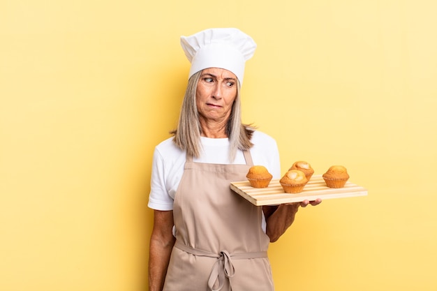 Donna chef di mezza età che si sente triste, sconvolta o arrabbiata e guarda di lato con un atteggiamento negativo, accigliata in disaccordo e con in mano un vassoio di muffin