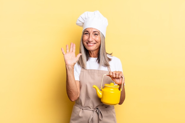 Donna chef di mezza età che sorride allegramente e allegramente, agitando la mano, accogliendoti e salutandoti, o salutandoti e tenendo una teiera