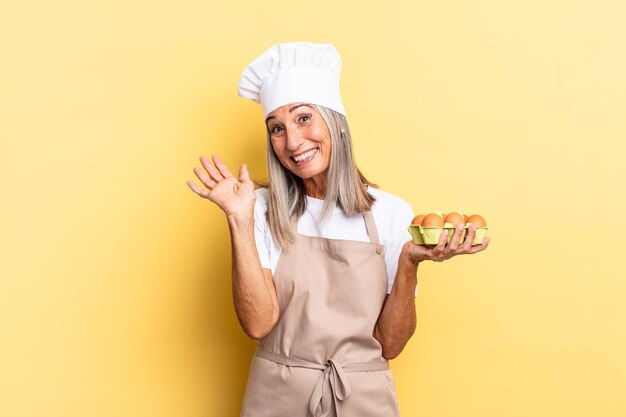 Donna chef di mezza età che sorride allegramente e allegramente agitando la mano dandoti il benvenuto e salutandoti o salutandoti tenendo una scatola di uova