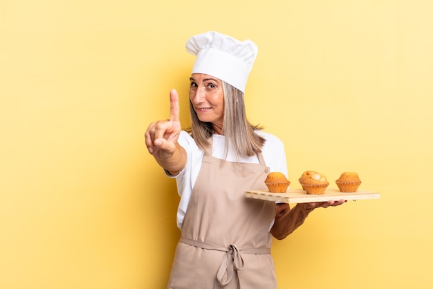 Donna chef di mezza età che sorride con orgoglio e sicurezza facendo la posa numero uno in modo trionfante, sentendosi come un leader e tenendo in mano un vassoio di muffin