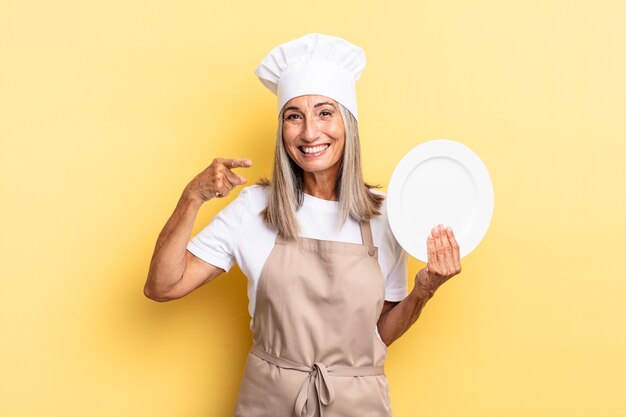 Donna chef di mezza età che sorride con sicurezza indicando il proprio ampio sorriso, atteggiamento positivo, rilassato, soddisfatto e tenendo in mano un piatto