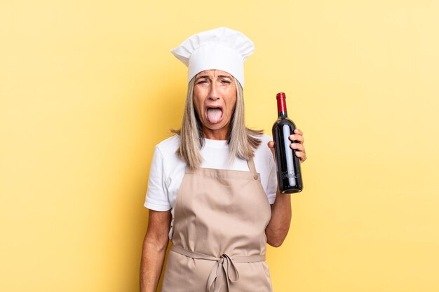 Donna chef di mezza età con atteggiamento allegro, spensierato e ribelle, scherzando e tirando fuori la lingua, divertendosi con una bottiglia di vino