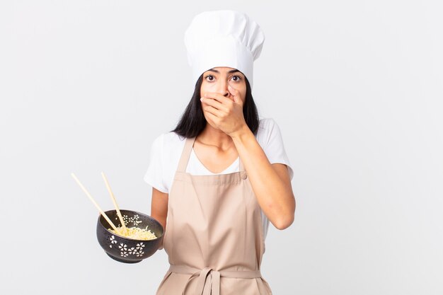 Donna chef piuttosto ispanica che copre la bocca con le mani con uno shock e tiene in mano una ciotola di noodle
