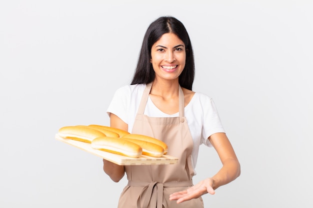 Donna chef piuttosto ispanica che sorride allegramente, si sente felice e mostra un concetto e tiene in mano un vassoio con focacce di pane