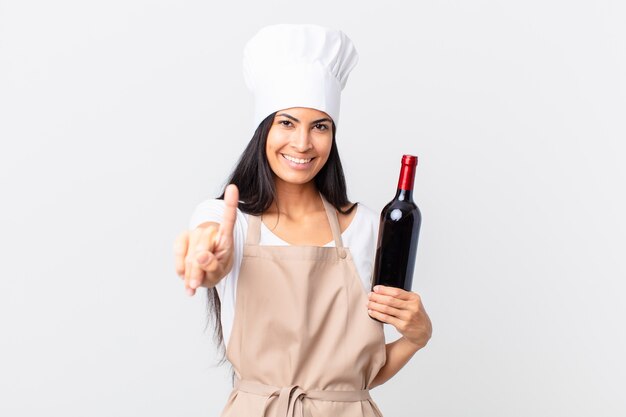 Donna chef piuttosto ispanica che sorride con orgoglio e sicurezza facendo il numero uno e tenendo in mano una bottiglia di vino