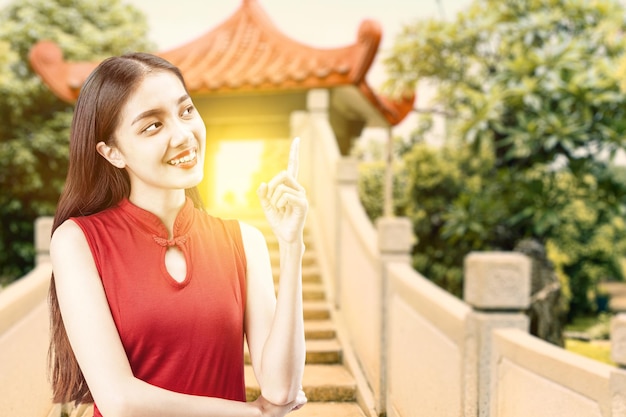 Donna cinese asiatica in un vestito cheongsam che indica qualcosa con il fondo cinese dell'edificio del gazebo. Buon Capodanno cinese