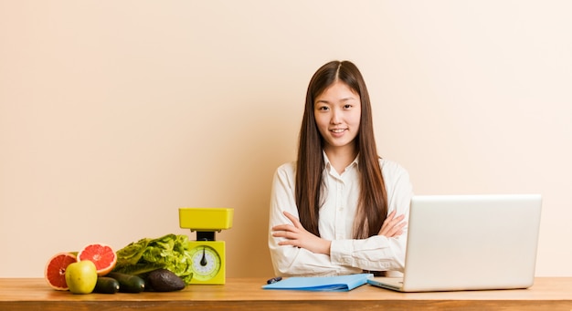 Donna cinese del giovane dietista che lavora con il suo sorridere del computer portatile sicuro con le braccia attraversate.