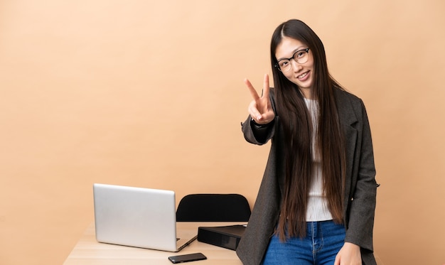 Donna cinese di affari nel suo posto di lavoro che sorride e che mostra il segno di vittoria