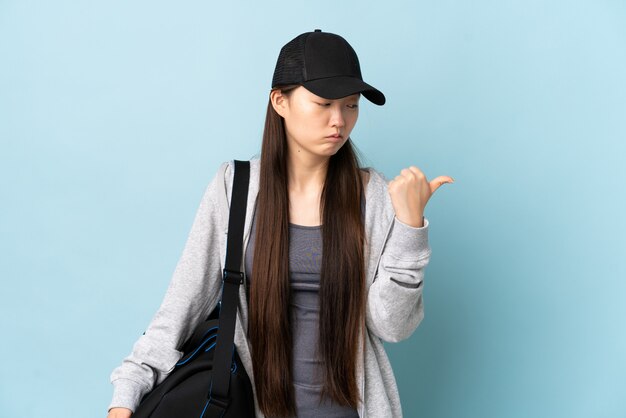 Donna cinese di giovane sport con la borsa di sport sopra la parete blu infelice e indicando il lato