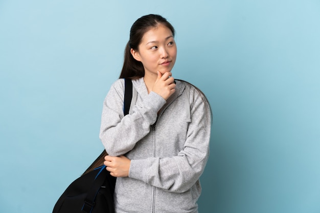 Donna cinese di giovane sport con la borsa di sport sopra la parete blu isolata che pensa un'idea mentre guardando su