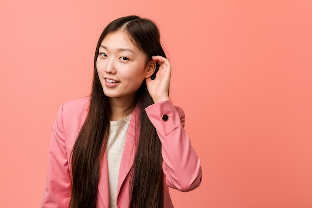 Donna cinese di giovani affari che indossa vestito rosa che prova ad ascoltare un gossip.