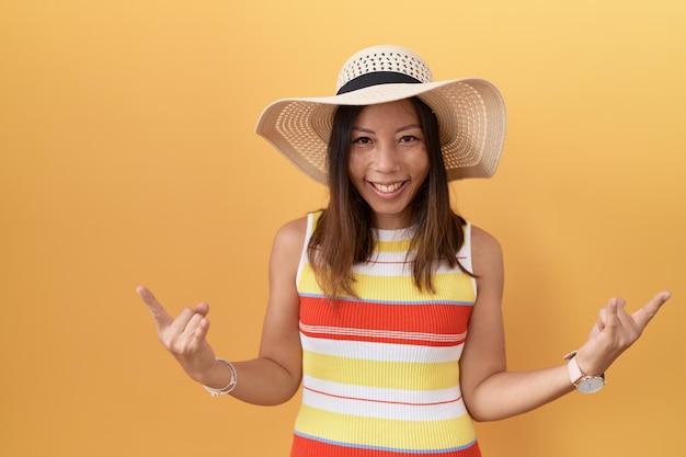 Donna cinese di mezza età che indossa cappello estivo su sfondo giallo che grida con un'espressione folle facendo il simbolo rock con le mani in alto concetto di musica pesante star della musica