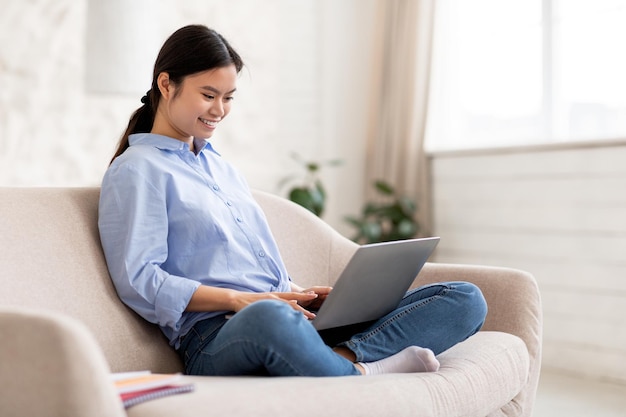 Donna cinese felice che scrive sull'interno domestico della tastiera del computer portatile