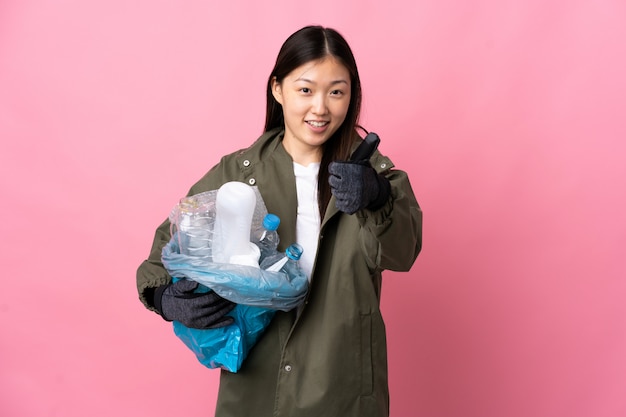 Donna cinese in possesso di un sacchetto pieno di bottiglie di plastica per riciclare isolato muro rosa con il pollice in alto perché è successo qualcosa di buono