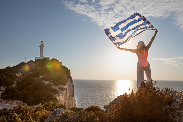 Donna con bandiera greca guardando il tramonto sopra il mare Faro dell'isola di Lefkada sullo sfondo