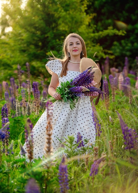 donna con bouquet di lupini in abito e cappello di paglia cammina sul prato con lupini al tramonto