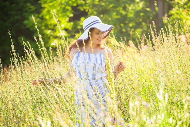 Donna con capelli lunghi in cappello e vestito che cammina attraverso la foresta di estate in una giornata di sole