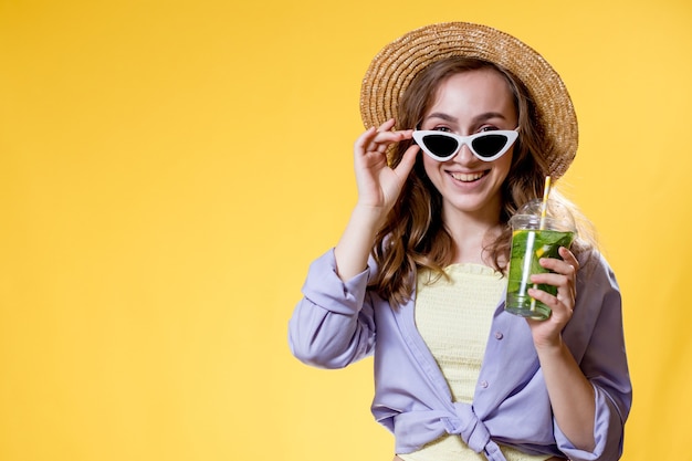 Donna con cappello di paglia che tiene gli occhiali da sole bianchi e il mojito in una tazza di plastica su sfondo giallo yellow