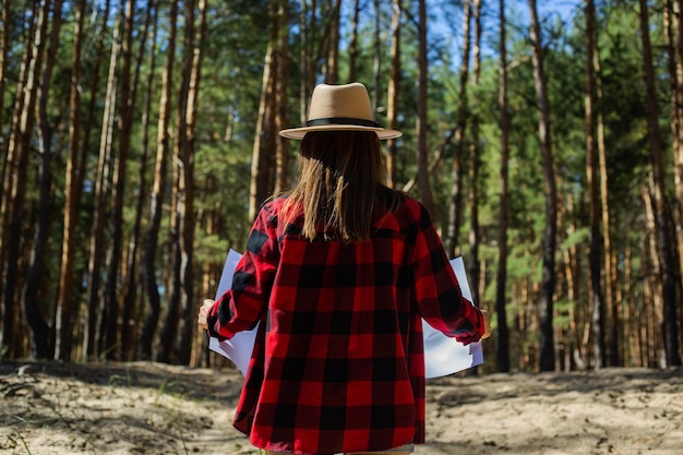 Donna con cappello e camicia a quadri rossa che tiene una mappa nella foresta