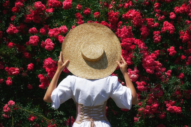 Donna con cappello e vestito bianco con corsetto cammina nel giardino sullo sfondo di cespugli di rose bella ragazza romantica