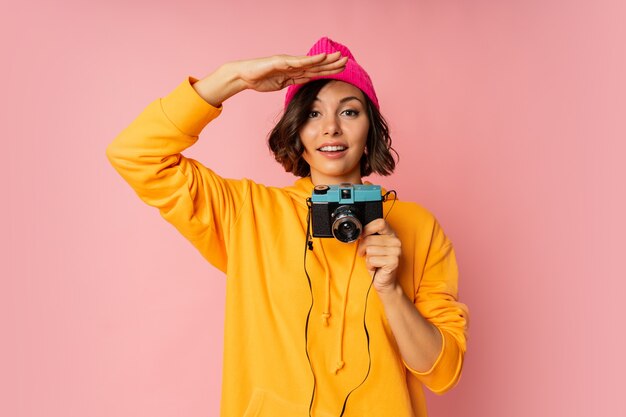 Donna con cappello rosa e felpa con cappuccio arancione con faccia suprice usando la macchina fotografica su rosa.