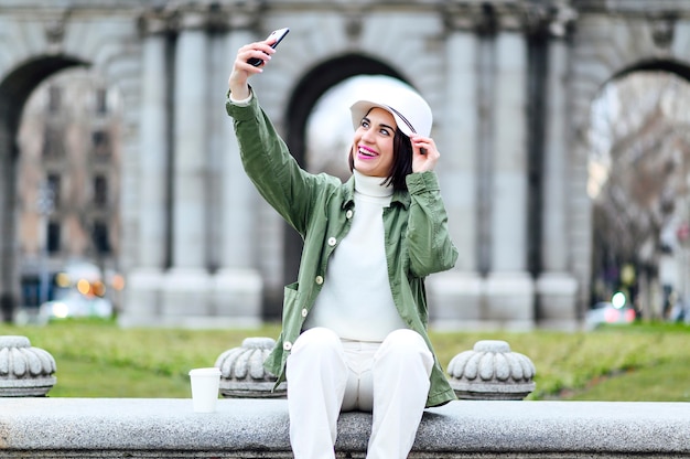 Donna con cappello seduta che si fa un selfie in città