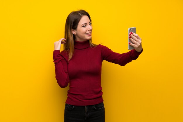 Donna con collo alto sopra muro giallo facendo un selfie