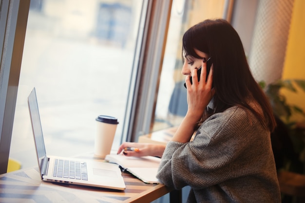 Donna con computer portatile e telefono cellulare che lavora in un caffè