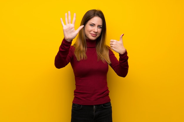 Donna con dolcevita su muro giallo contando sei con le dita